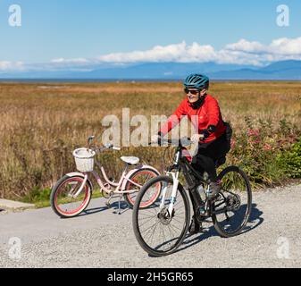 Sorridente donna anziana in bicicletta in un parco. Donna anziana felice che indossa il casco sulla moto sportiva. Foto di strada, foto di viaggio, invecchiamento attivo Foto Stock