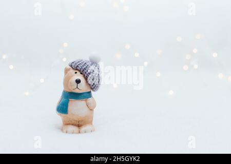 Figurine decorative a tema natalizio. La statuetta di un orso in un cappello a maglia su sfondo bianco. Decorazioni festive, calde luci bokeh Foto Stock