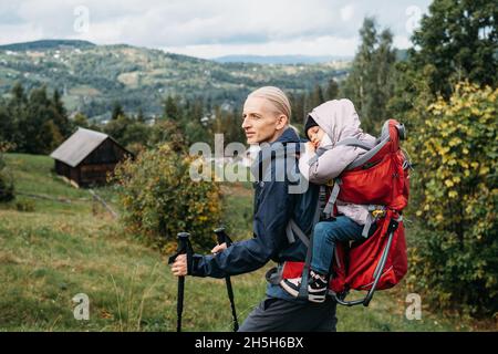 Uomo viaggiatore con bambino stanco in zaino Padre porta sua figlia in uno zaino Male ha preso bambino scivolante verso le montagne. Viaggiare con bambini Foto Stock