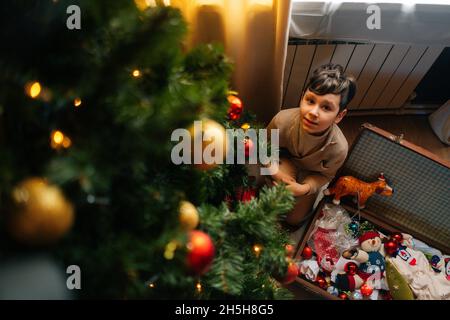 Vista ad angolo di adorabile bambino ragazzo decorazione albero di Natale con palle lucenti e giocattoli seduti sul pavimento con vecchia valigia di famiglia giocattoli di Natale. Foto Stock