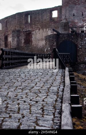 Strada acciottolata e ponte levatoio in legno con cancello chiuso e mura in rovina del castello in background primo piano rovine del castello dei vescovi silesiani Siewierz Foto Stock