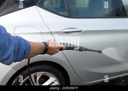 primo piano della mano di mans, tenendo un ugello di lavaggio a pressione, lavando a mano una vettura argentata all'esterno sul vialetto in una giornata di sole Foto Stock