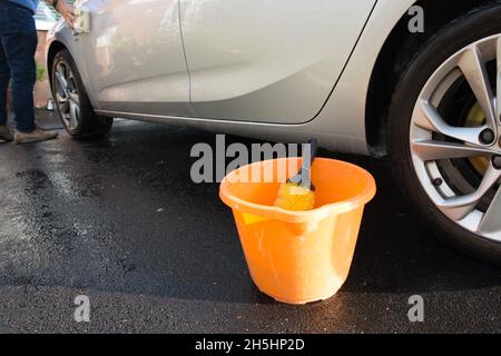 primo piano del secchio arancione e della spazzola di lavaggio auto con un'auto d'argento su strada asfaltata all'esterno in una giornata di sole Foto Stock