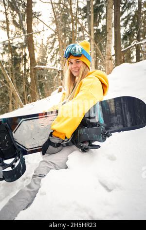 Donna caucasica snowboarder tiene snowboard sullo sfondo di una bella foresta innevata in giornata di sole Foto Stock