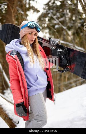 Donna caucasica snowboarder tiene snowboard sullo sfondo di una bella foresta innevata in giornata di sole Foto Stock