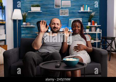 Coppia interrazziale in attesa del bambino e utilizzando la videochiamata per la comunicazione remota online a casa. Genitori multietnici che ondeggiavano mentre guardavano la macchina fotografica per conferenze via internet Foto Stock