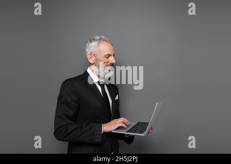 uomo d'affari felice di mezza età in tuta usando il laptop in grigio Foto Stock