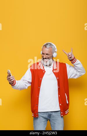 positivo uomo di mezza età in cuffie prendendo selfie mentre mostra rock segno sul giallo Foto Stock