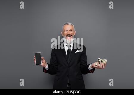 gioioso uomo d'affari di mezza età che detiene banconote in dollari e smartphone con schermo vuoto in grigio Foto Stock