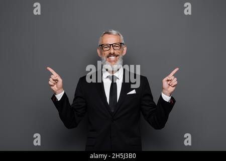 uomo d'affari positivo di mezza età in occhiali puntando con le dita a parte su grigio Foto Stock