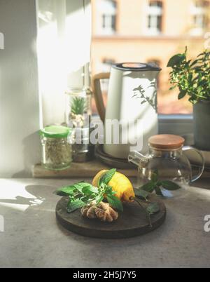 Preparazione di tè allo zenzero fresco con ingredienti sani a casa. Zenzero, foglie di menta e limone sul tavolo da cucina con tea pot in vetro, bollitore elettrico e ja Foto Stock