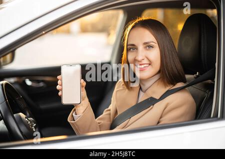 La giovane donna felice si siede all'interno della nuova auto moderna, tiene lo smartphone con schermo vuoto, mostrando la nuova app mobile per i conducenti, spazio copia Foto Stock