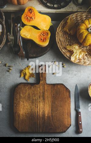 Fondo del cibo con tagliere in legno vuoto sul tavolo da cucina con zucca a metà, posate e utensili da cucina. Scena di cucina autunnale con Foto Stock