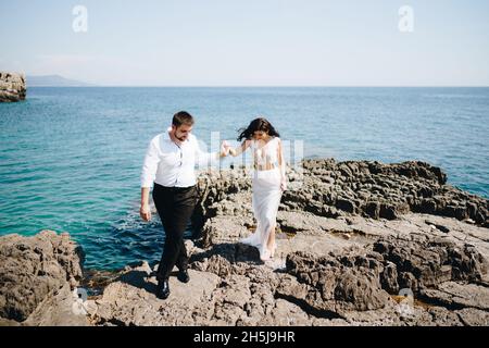 La sposa e lo sposo camminano tenendo le mani sulla riva rocciosa Foto Stock