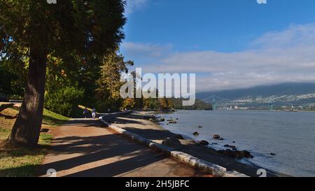 Splendida vista delle persone che si godono una passeggiata al Seawall Trail in Stanley Park, Vancouver, British Columbia, Canada con Lions Gate Bridge in background. Foto Stock