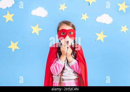 Il bambino gioca il supereroe. Capretto sullo sfondo di una parete blu brillante con nuvole bianche e stelle. Concetto di potere della ragazza. Foto Stock