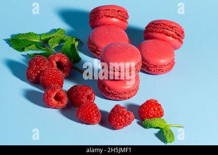 Un assortimento di vivaci macaron di lamponi rossi, accompagnati da lamponi freschi e foglie di menta, disposti su uno sfondo blu pastello Foto Stock