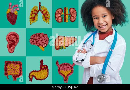 Concetto di salute dei bambini, pediatria. Sorridente bambina afroamericana in cappotto medico in piedi vicino a icone anatomiche con illustrazioni interne o Foto Stock