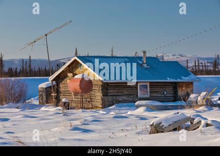 Una capanna di legno è circondata da neve, antenna TV e tamburo di olio di riscaldamento in inverno, Nulato, interno Alaska, USA; Nulato, Alaska, Stati Uniti d'America Foto Stock