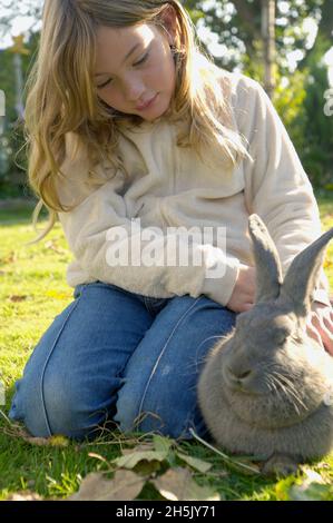 Ragazza che si inginocchia sull'erba accarezzando un coniglio grigio (Oryctolagus cuniculus domesticus); Germania Foto Stock