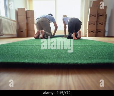 Vista presa da dietro di coppia che si inginocchia sul pavimento che arrotola il tappeto in una nuova casa tra pile di scatole di cartone; Germania Foto Stock