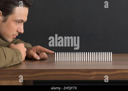 Primo piano di un uomo che si prepara a spingere il primo domino in una fila di dominazioni su un tavolo, anticipando i risultati; Germania Foto Stock