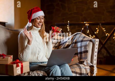 Concetto di festa virtuale. Eccitata ragazza allegra in Babbo Natale Hat Holding Gift Box con regalo di Natale, seduto sul divano in salotto, avendo Vid Foto Stock