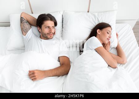 Deluso triste giovane caucasico ragazzo non dorme, soffre di rumore guarda la moglie sul letto in camera da letto interno. Russare, apnea, insonnia, litigio e. Foto Stock