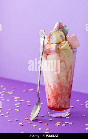 Tazza di vetro con frullato e sciroppo di frutta con confetti di stelle, marshmallow multicolore e cucchiaio su sfondo viola brillante, concetto di compleanno Foto Stock