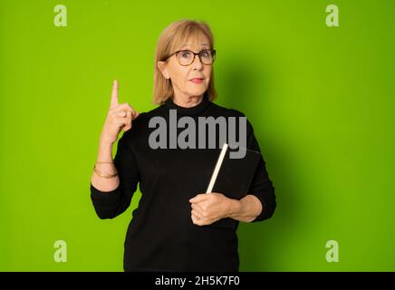 Immagine di una donna anziana che tiene un libro puntando il dito su isolato su sfondo verde. Foto Stock