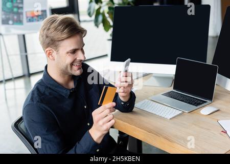 Positivo sviluppatore che utilizza smartphone e carta di credito vicino a computer in ufficio Foto Stock
