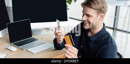 Programmatore sorridente con smartphone e carta di credito vicino a computer in ufficio, banner Foto Stock
