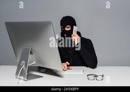 Hacker in balaclava parlando su smartphone e utilizzando il computer vicino occhiali isolati in grigio Foto Stock