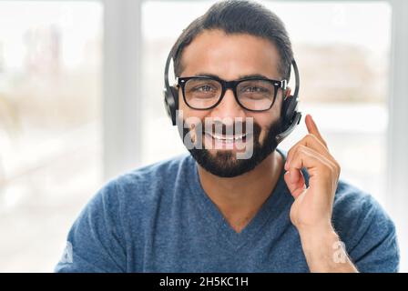 Addetto all'assistenza clienti che indossa un auricolare. Fidato uomo indiano dipendente in cuffie con microfono guarda la fotocamera con sorriso amichevole. Chat video con un operatore di call center orientale Foto Stock