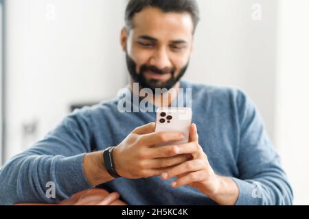 Fuoco selettivo allo smartphone trendy nelle mani del ragazzo indiano concentrato giovane, il freelancer maschio ispanico che usa il telefono per il lavoro remoto dalla casa, uomo etnico che usa la nuova app mobile, inviare SMS online Foto Stock