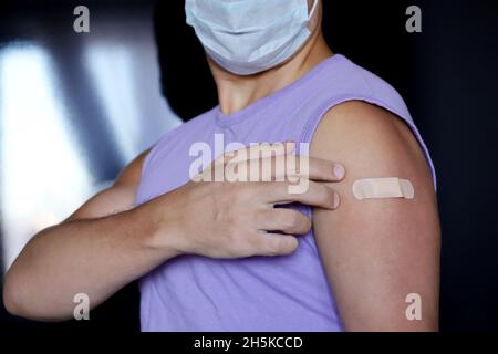 Uomo in maschera protettiva viso che mostra braccio con bendaggio dopo aver ricevuto il vaccino. Vaccinazione durante la pandemia di covid-19 Foto Stock