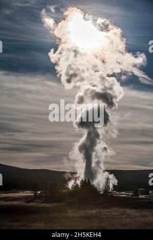 Sole che illumina una colonna di vapore termale che si diffonde da Old Faithful erutting contro un cielo nuvoloso, blu e sagome cresta di montagna in The Up... Foto Stock