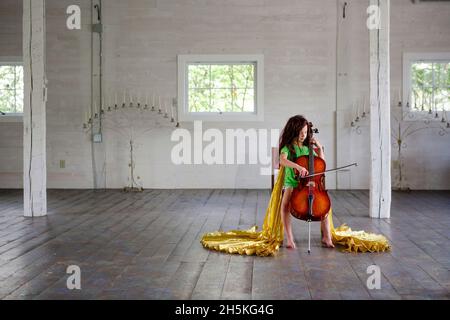 La ragazza Barefoot in vimini e mantello d'oro suona violoncello in un grande fienile vuoto Foto Stock