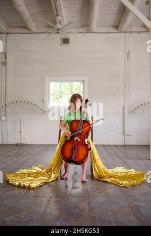 Un bambino piccolo con lunghe ali d'oro gioca violoncello da solo in fienile vuoto Foto Stock