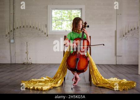 Una bambina in lunghe ali dorate suona violoncello in un fienile vuoto Foto Stock