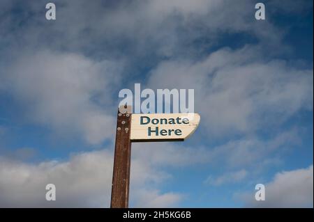 Donare qui segno su legno dipinto di bianco, su palo di legno scuro. Isolato contro cielo blu e sfondo nuvole. Molto spazio per le copie. Foto Stock