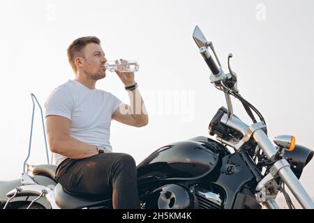 Un ragazzo giovane e attraente siede su una moto e beve acqua da una bottiglia. Un bel giovane uomo in una T-shirt bianca si siede e si appoggia su una bicicletta. Sole caldo Foto Stock