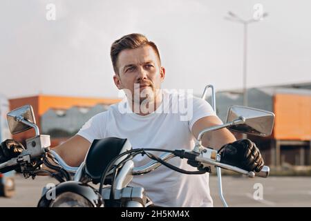 Elegante e accattivante giovane uomo in T-shirt bianca si siede su una motocicletta. Ritratto bel motociclista in posa su una bicicletta. Fotografia di stile di vita. Sfondo urbano Foto Stock