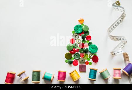 Un albero di Natale fatto di bottoni di colore verde, rosso. Fili multicolore con bobine in legno, nastro di misurazione. Sfondo bianco, spazio di copia. Chr. Creativa Foto Stock
