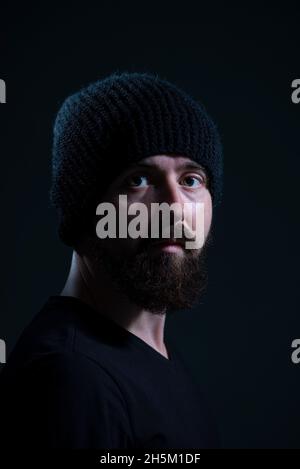 autentico ritratto drammatico di un millenario con una barba in un cappello di maglia nero, su sfondo scuro. Foto Stock