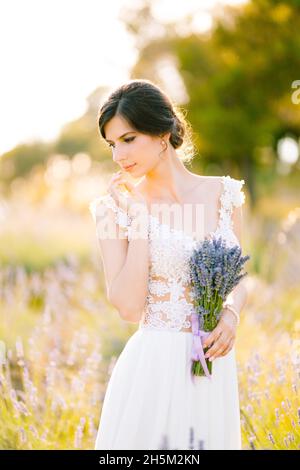 Sibenik, Croazia - 05.06.17: Sposa in un abito in pizzo bianco si erge in un campo e tiene in mano un bouquet di lavanda legato con un nastro di lilla, girando Foto Stock