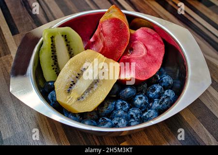 in una ciotola di acciaio inossidabile su un tavolo, ha dimezzato i kiwi verdi e dorati, i mirtilli e una mela chiamata luna rossa Foto Stock