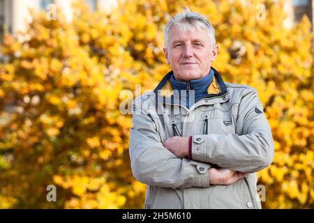 L'anziano caucasico con le braccia ripiegate sul petto, foglie di albero giallo come sfondo, stagione autunnale. CopySpace a sinistra Foto Stock
