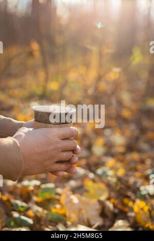 Un uomo tiene una tazza di carta usa e getta con una bevanda nel parco in autunno. Il concetto di bevande, tè, caffè, autunno, riscaldamento, riposo. Foto Stock