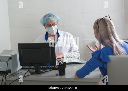 Un medico in un cappotto bianco, maschera e cappuccio è seduto a un tavolo con un paziente in ufficio e discutere il protocollo di trattamento. Ricettio consultivo Foto Stock